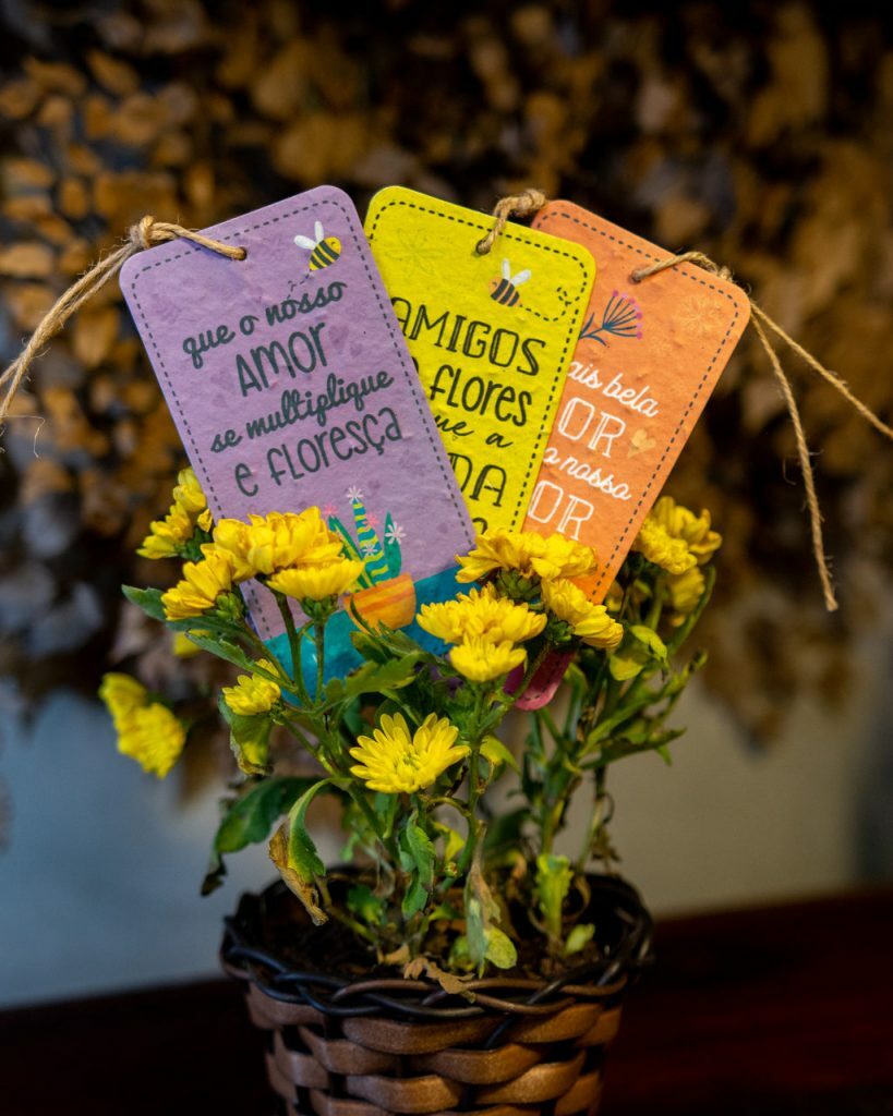 Tag-atividades-meio-ambiente. Um vaso com flores amarelas e três tags coloridas com mensagens inspiracionais.